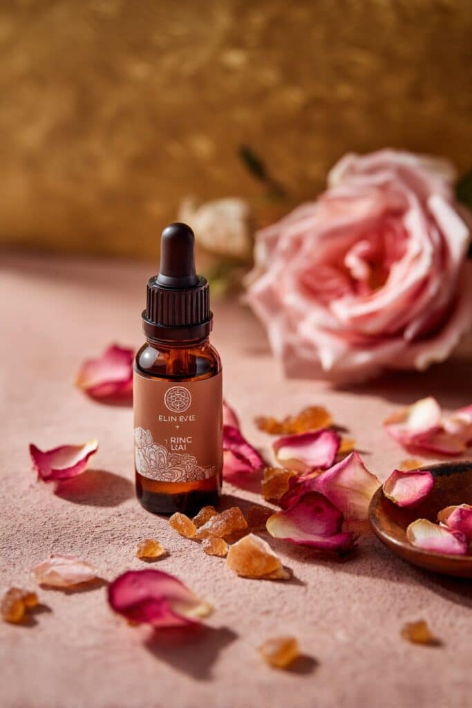 Amber dropper bottle containing anti-aging blend, surrounded by rose petals and frankincense resin on muted pink and golden beige background.