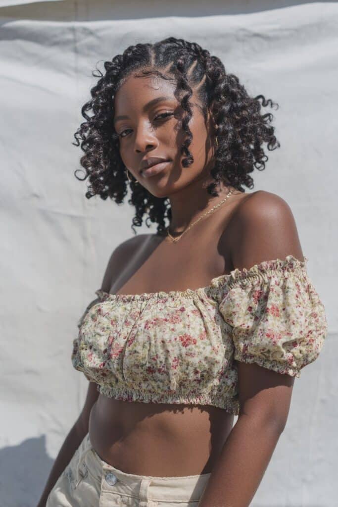 Stylish young American woman with curly hair in bubble braids, wearing floral off-shoulder blouse with carefree expression against white backdrop.