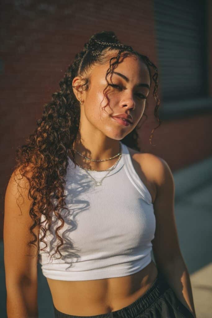 Young American woman with curly hair in tight Dutch braids, wearing sporty crop top and biker shorts against urban brick wall.