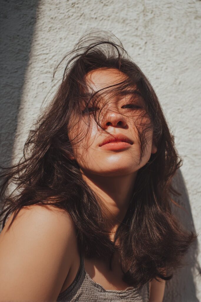 American woman with ghost layers in medium-length hair, airy style, dewy makeup, summery top, plaster backdrop.