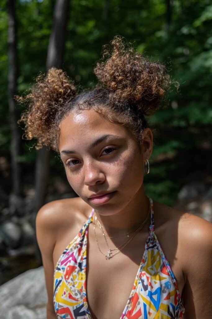 Trendy young American woman with curly hair in half-up space buns, wearing colorful halter top in natural forest setting.
