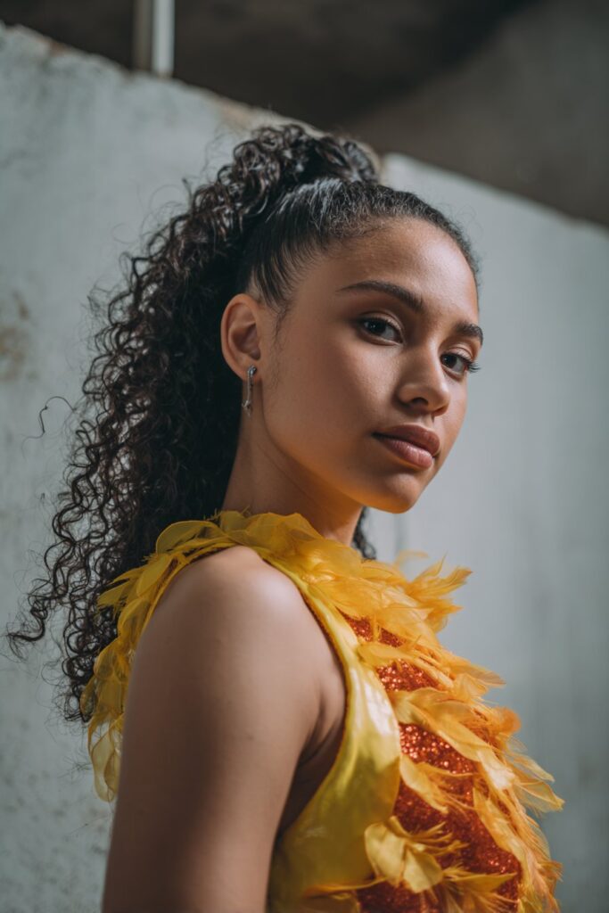 American woman with curly hair in high sleek ponytail, vibrant dance costume, spotlight-ready pose, textured plaster backdrop.