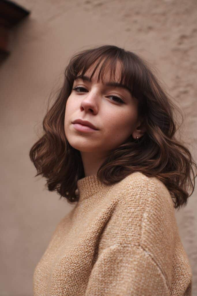 American woman with chin-length bangs and soft long layers, natural waves, cozy autumn outfit against textured plaster backdrop.
