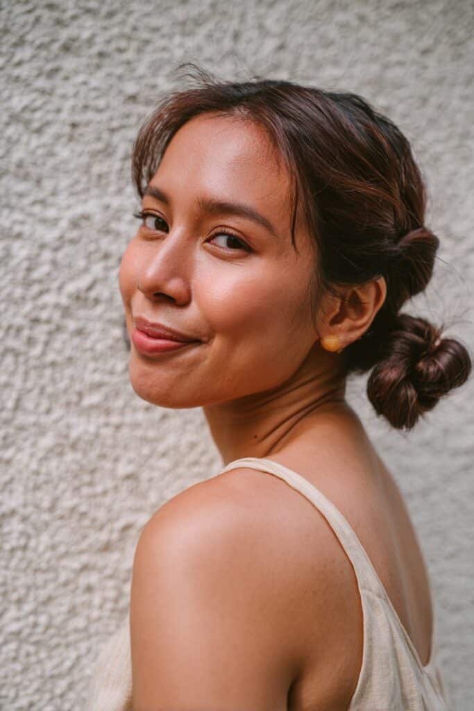 American woman with hair in two low twisted buns, sleeveless top, soft smile, textured plaster wall background.