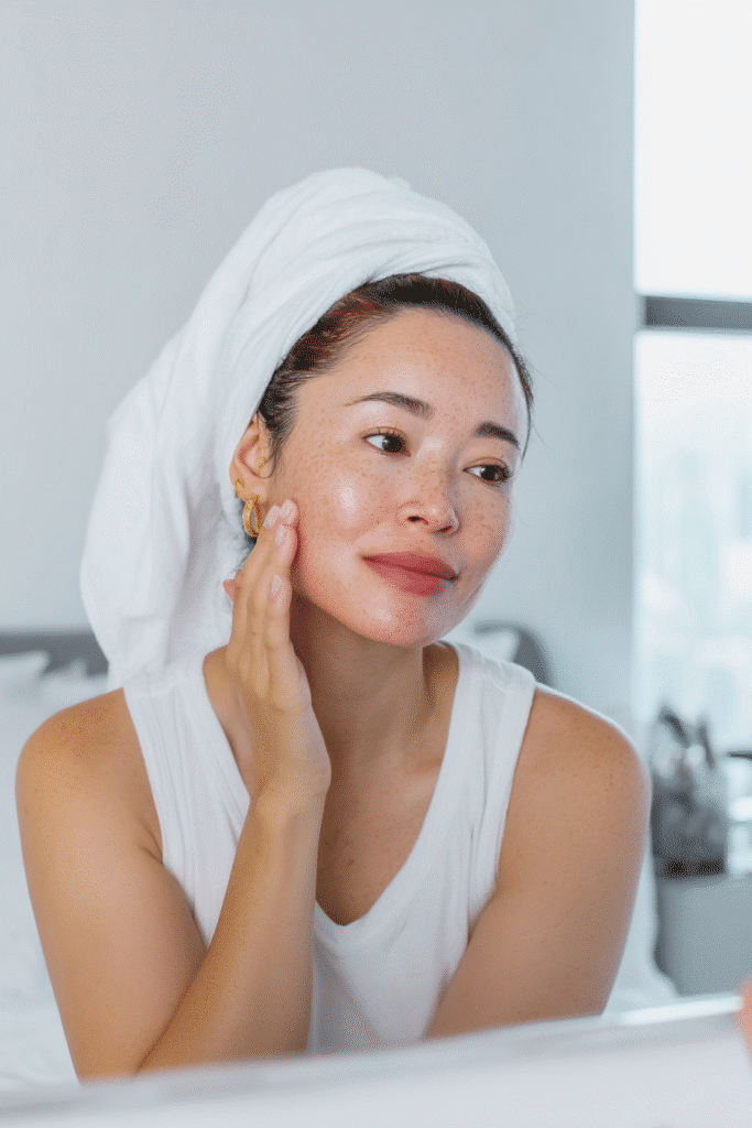 Woman with towel-wrapped hair applying facial moisturizer, looking in bathroom mirror with natural lighting.