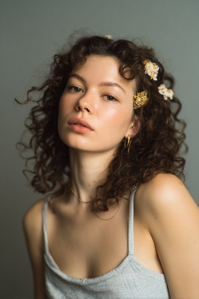 Young American woman with curly hair styled with gold bobby pins, wearing spaghetti strap top with summer glow against gray fabric