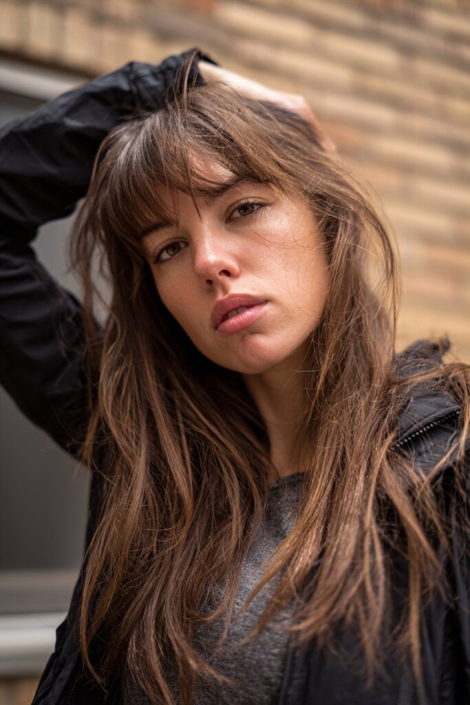 American woman with messy shag haircut, peek-a-boo bangs, playful expression, casual street style clothing against urban brick wall background.