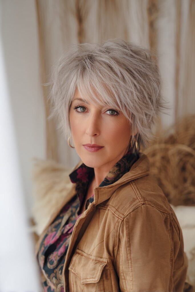 "Sophisticated portrait of mature woman with silver face-framing shag in neutral boho interior with soft curtains."