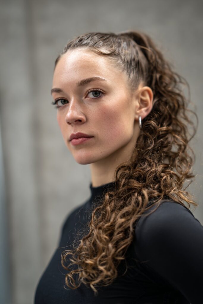 American woman with curly hair in slicked back bun, refined dance competition styling, focused expression, gray backdrop.