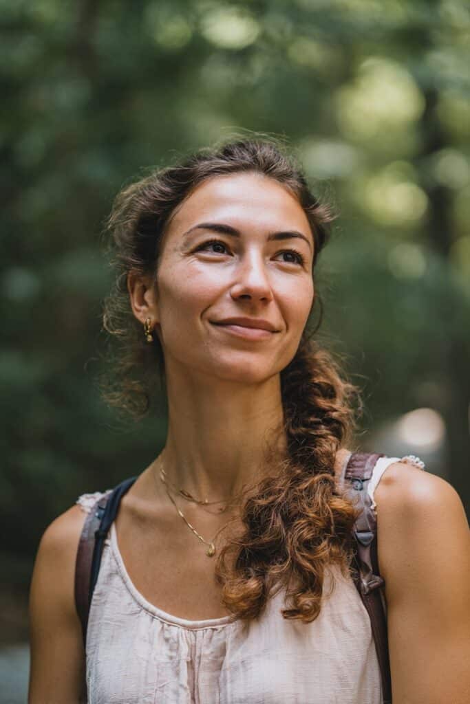 American woman with curly hair in side twisted braid, gentle smile, lightweight summer top, natural forest background.