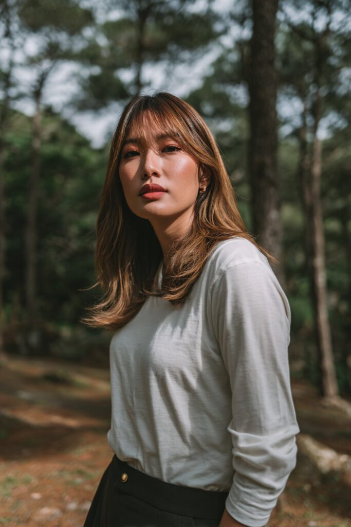 American woman with V-cut long layers and sweeping bangs, soft glam makeup, classy outfit against natural forest backdrop.