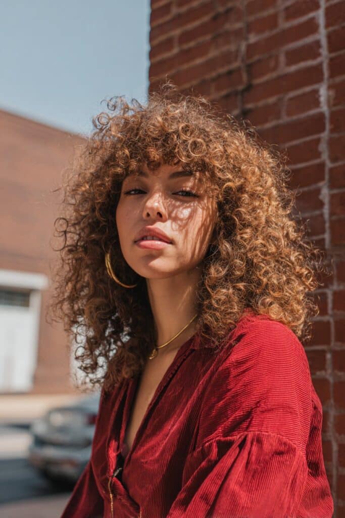 Retro-inspired portrait of American model with voluminous seventies shag, defined curtain bangs against urban brick wall background.