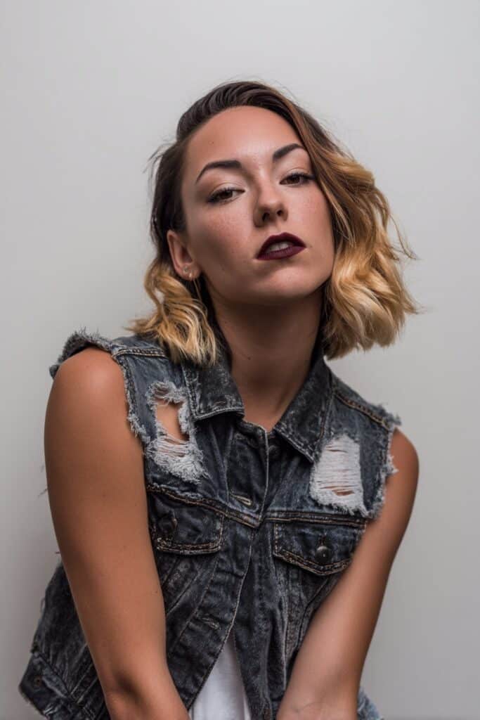 American female model with black shag featuring blonde money pieces, edgy emo glam, dark lipstick, denim vest, white backdrop.