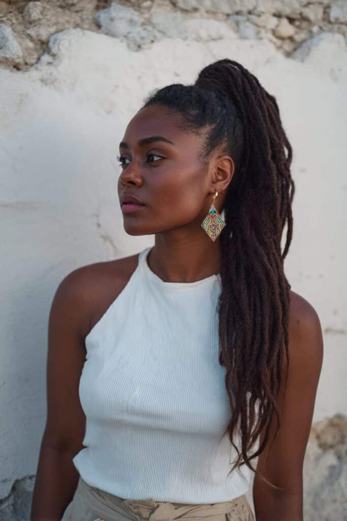 Fashion portrait of American woman with waist-length box braids in high ponytail wearing bold earrings against textured wall.