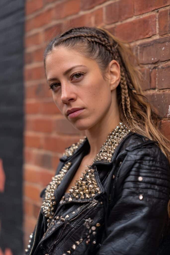 American woman with edgy braided side undercut, studded leather rocker jacket and bold attitude against urban brick wall.