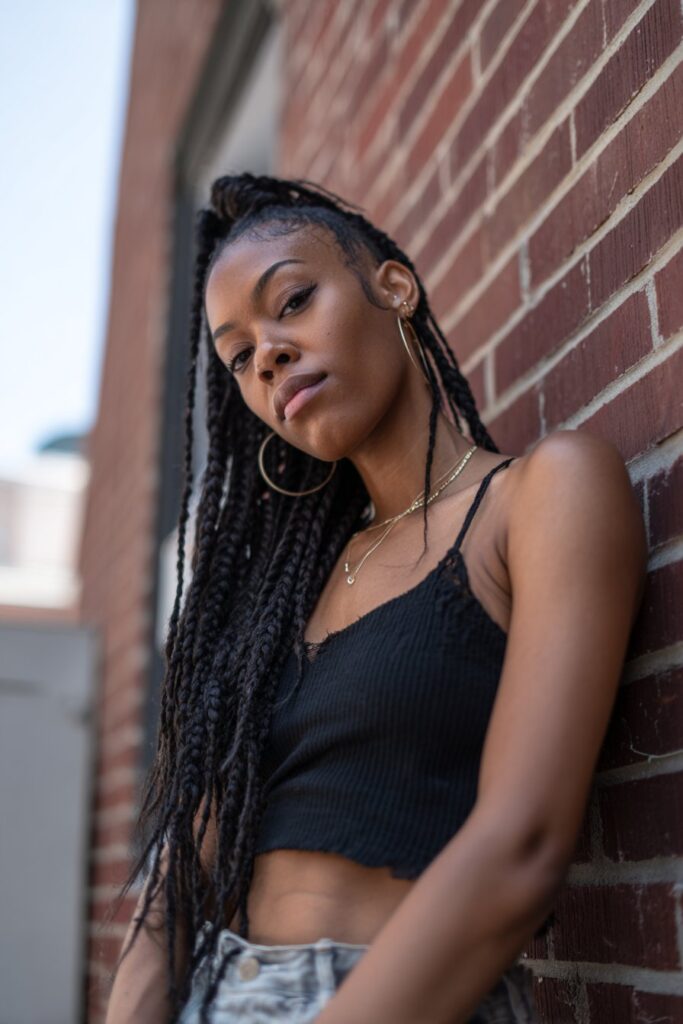 Portrait of American model with full braids and dramatic swooped baby hairs against urban brick wall backdrop.