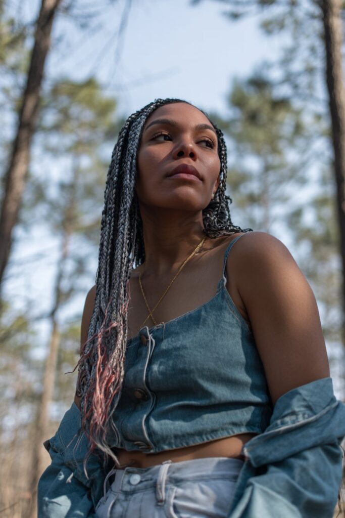 Outdoor portrait of American model with black braids featuring pink and copper highlights against natural forest background.