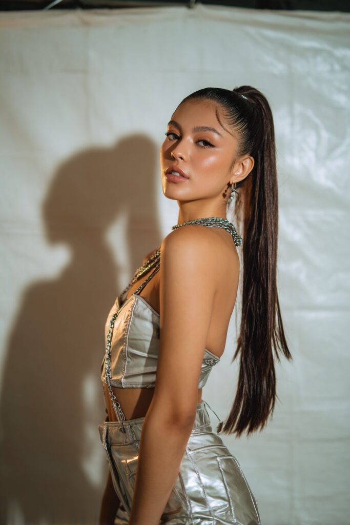 American woman with sleek bubble ponytail, metallic accessories, stage lighting, glamorous concert look on white backdrop.