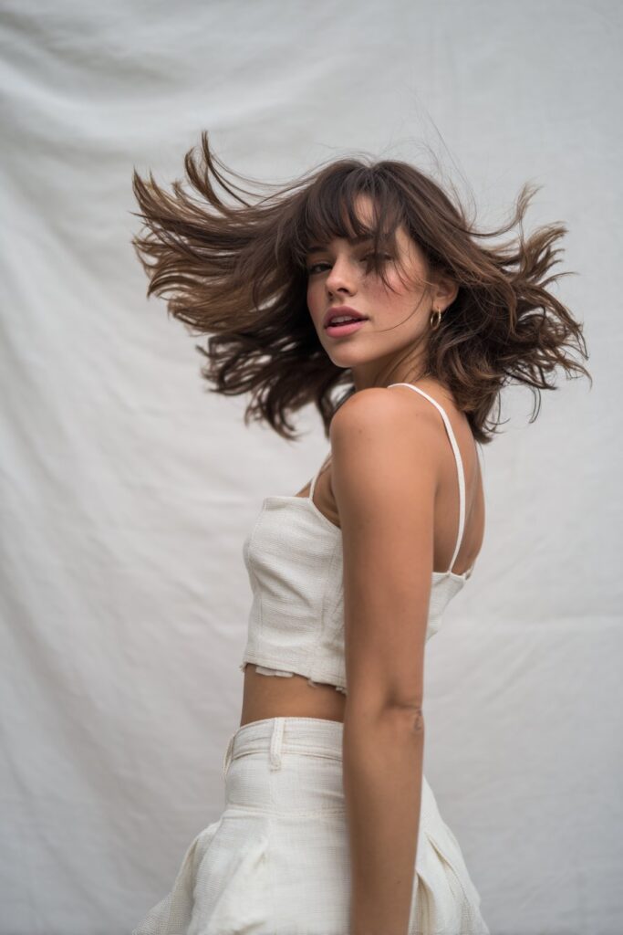 American woman with butterfly haircut, flippy ends, long side bangs, voluminous outward layers, playful expression, white backdrop.