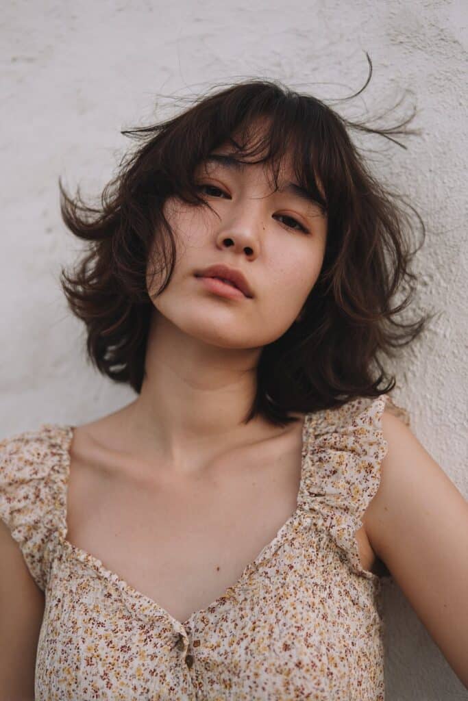 American model with butterfly haircut for fine hair, airy layers, front bangs, subtle volume, textured plaster backdrop.