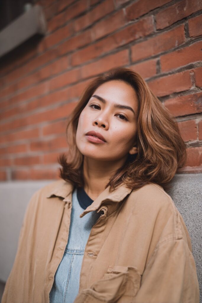 Portrait of American woman with round face and highlighted butterfly haircut wearing denim against brick wall.
