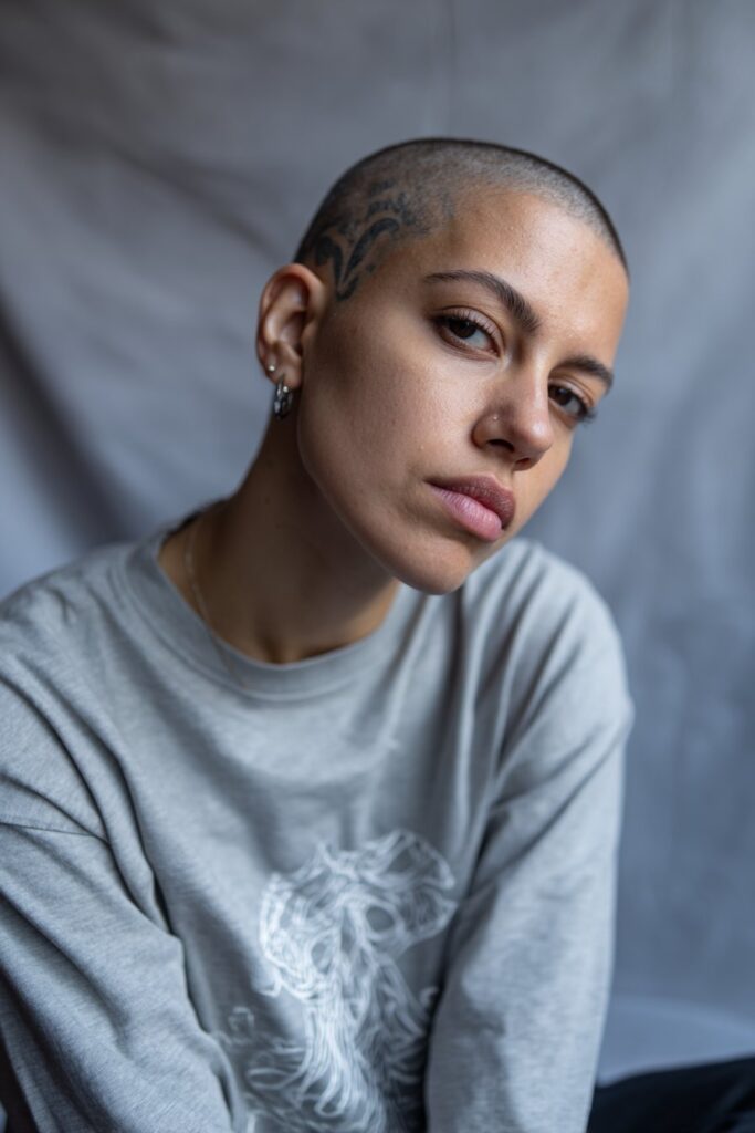 American female model with super short buzz cut featuring shaved design pattern, fierce expression, bold streetwear, gray backdrop.
