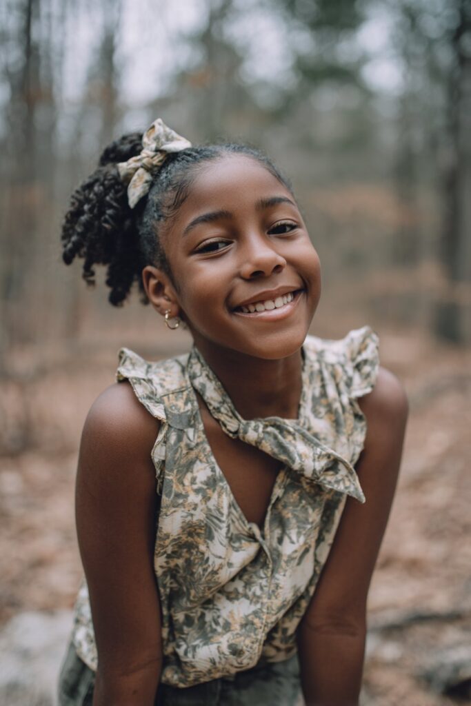 American girl with front cornrows in half updo, back curls free, pretty bow clip, joyful smile, natural forest.