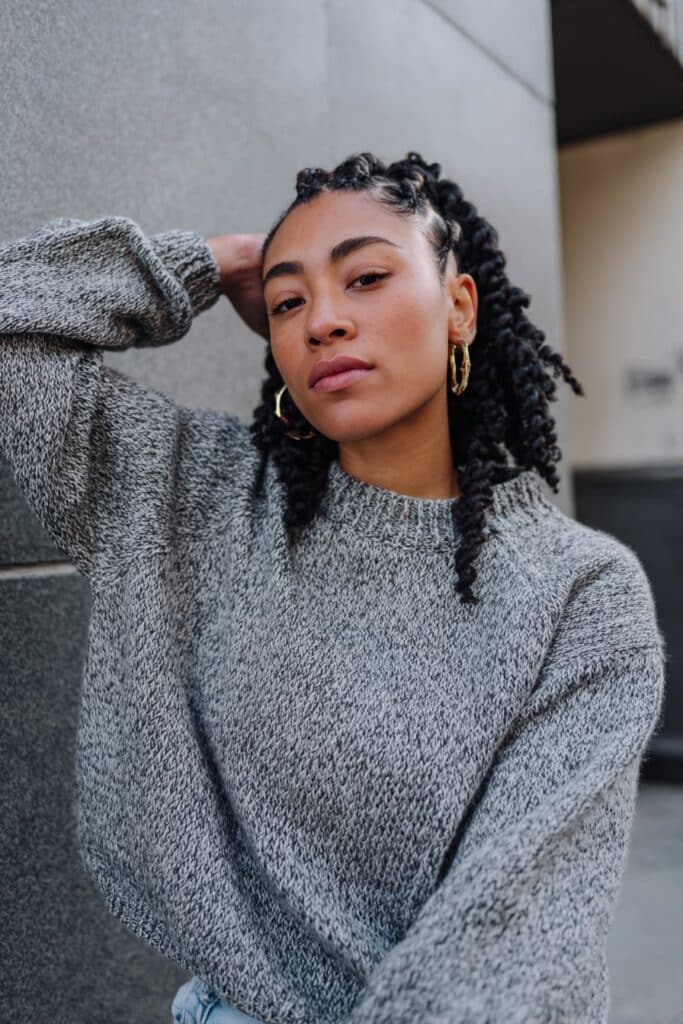Stylish Black woman with cornrows and voluminous crochet curls, modern streetwear, soft gray fabric backdrop.