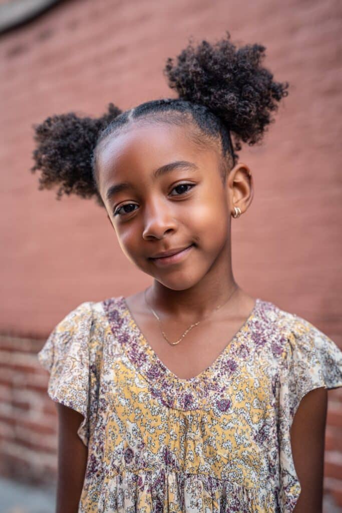 American girl with front cornrows leading to two high afro puffs, energetic style, bright patterned dress against brick wall.