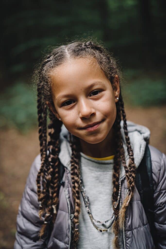 American kid with cornrows leading to twisted ends hanging freely, wearing playful accessories, relaxed expression, natural forest background.