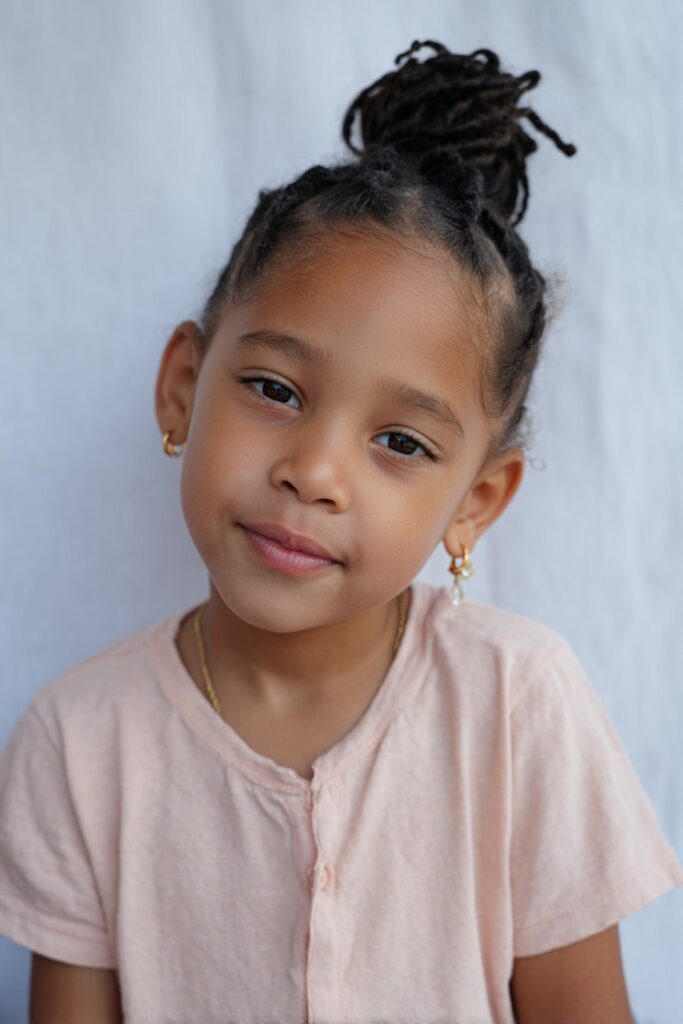 Young American child with neat cornrows in low bun, wearing cute earrings and pastel top, smiling against white backdrop.