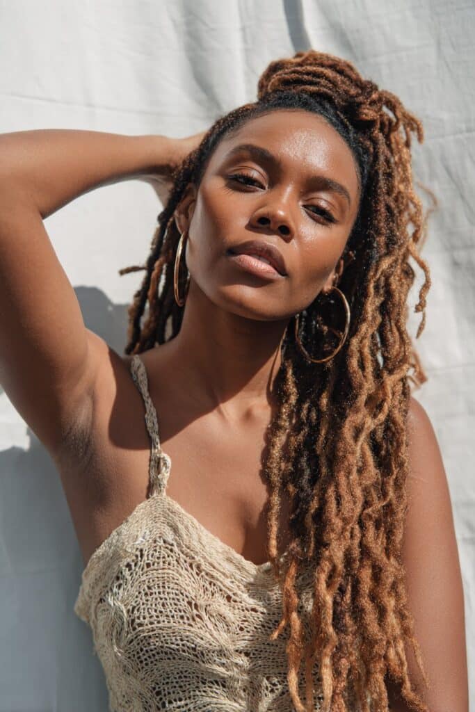 Black woman with long crochet boho locs, chestnut blonde highlights, hoop earrings, white seamless backdrop.