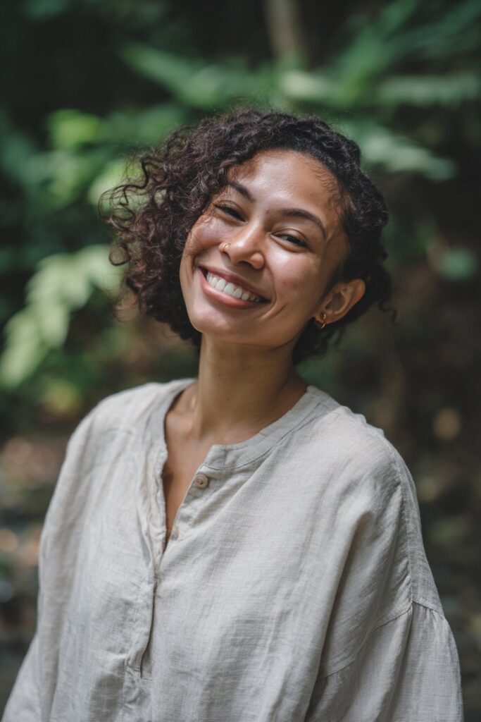 Smiling Black woman with spring twist crochet hair, shoulder-length coils, linen blouse, forest backdrop.