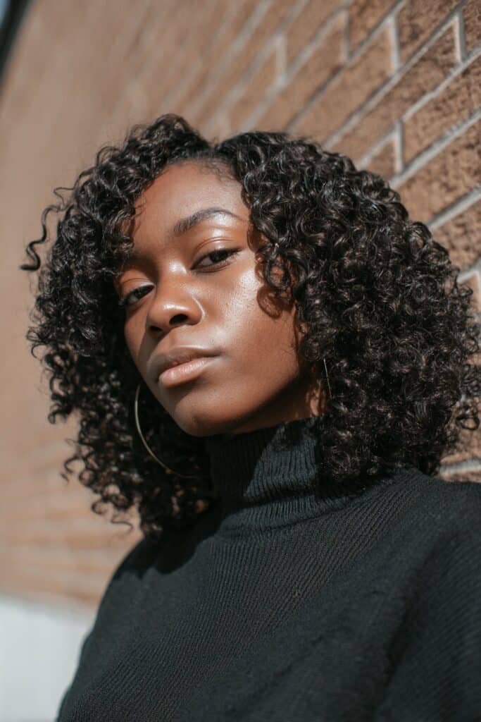 Elegant Black woman in crochet wig with tight curls, minimal makeup, black turtleneck, brick backdrop.