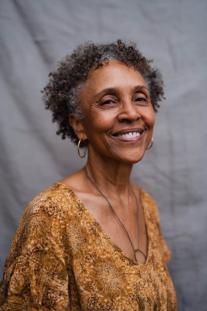Radiant older American woman with naturally curly hair, short curly bangs, big smile, vibrant top, soft gray fabric backdrop.