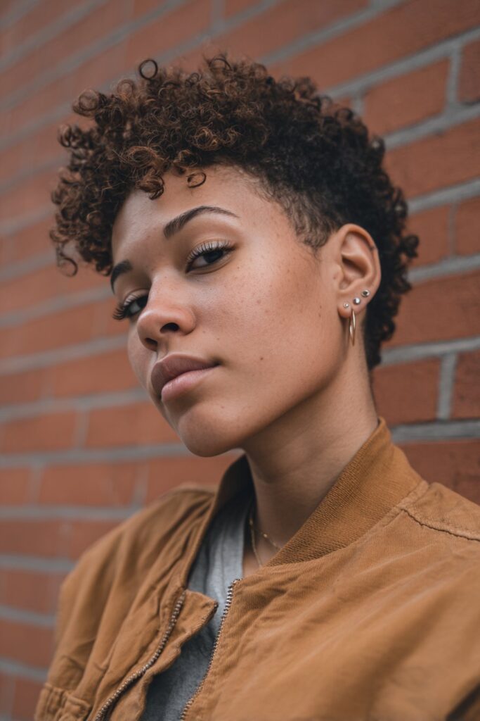 American female model with curly tapered short haircut, tight defined curls, warm skin tone, trendy urban outfit, brick wall.