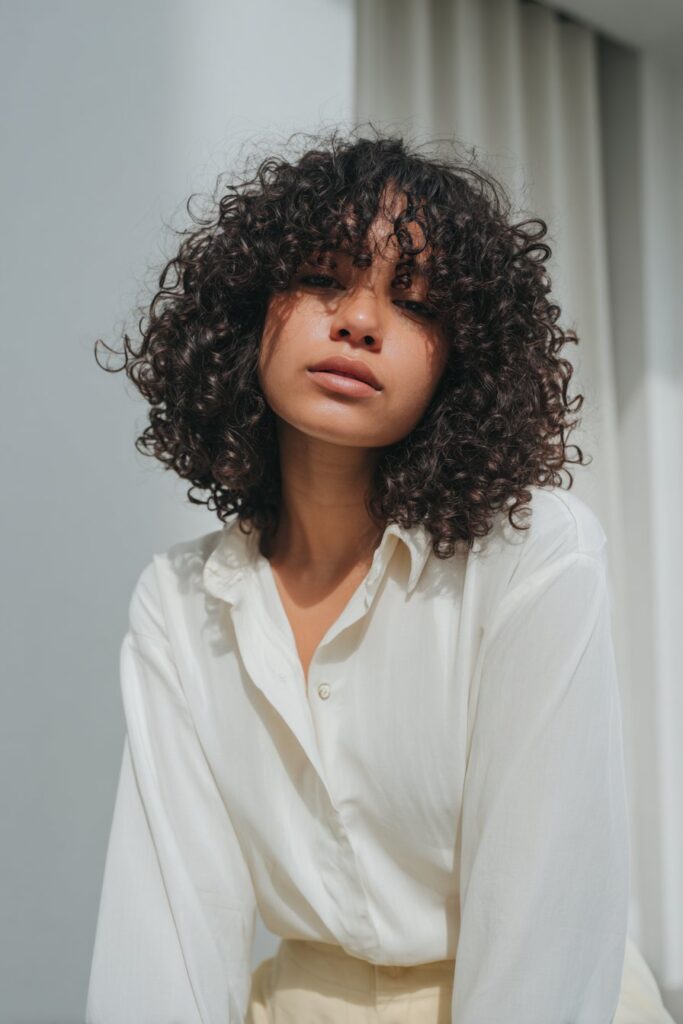Sleek portrait of American woman with stretched curls, curtain bangs, soft-toned outfit on white backdrop.