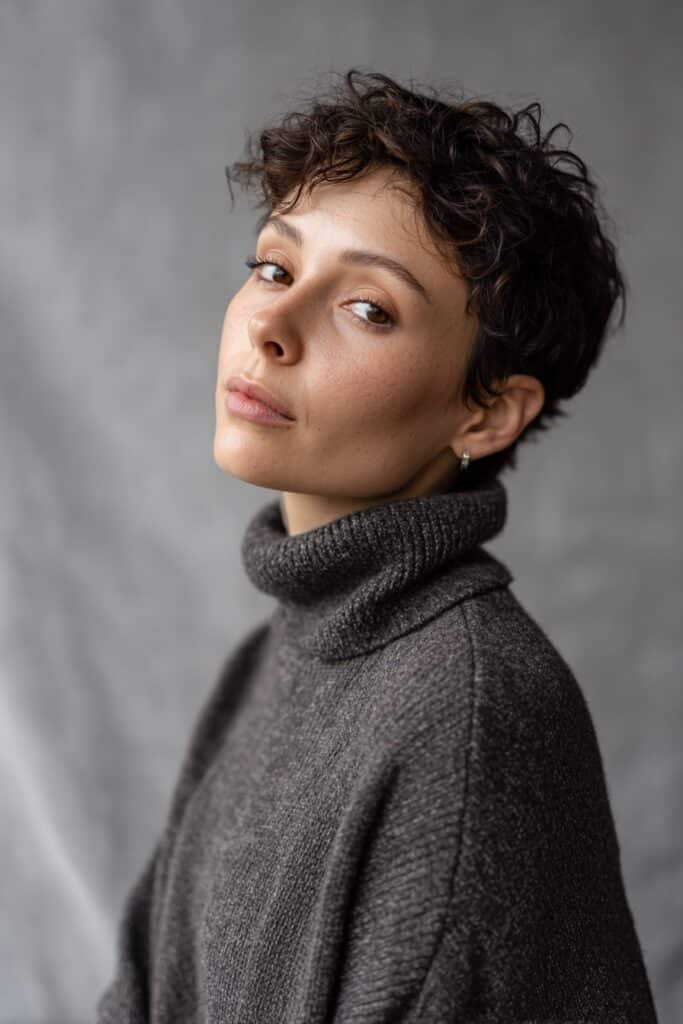 American woman with 4C hair pixie cut, dramatic deep side part, subtle makeup, soft natural light, gray fabric background.