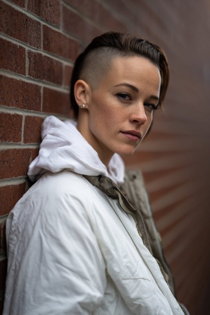American female model with disconnected undercut, longer slicked back top, clean shaved sides, edgy makeup, modern streetwear, brick wall.