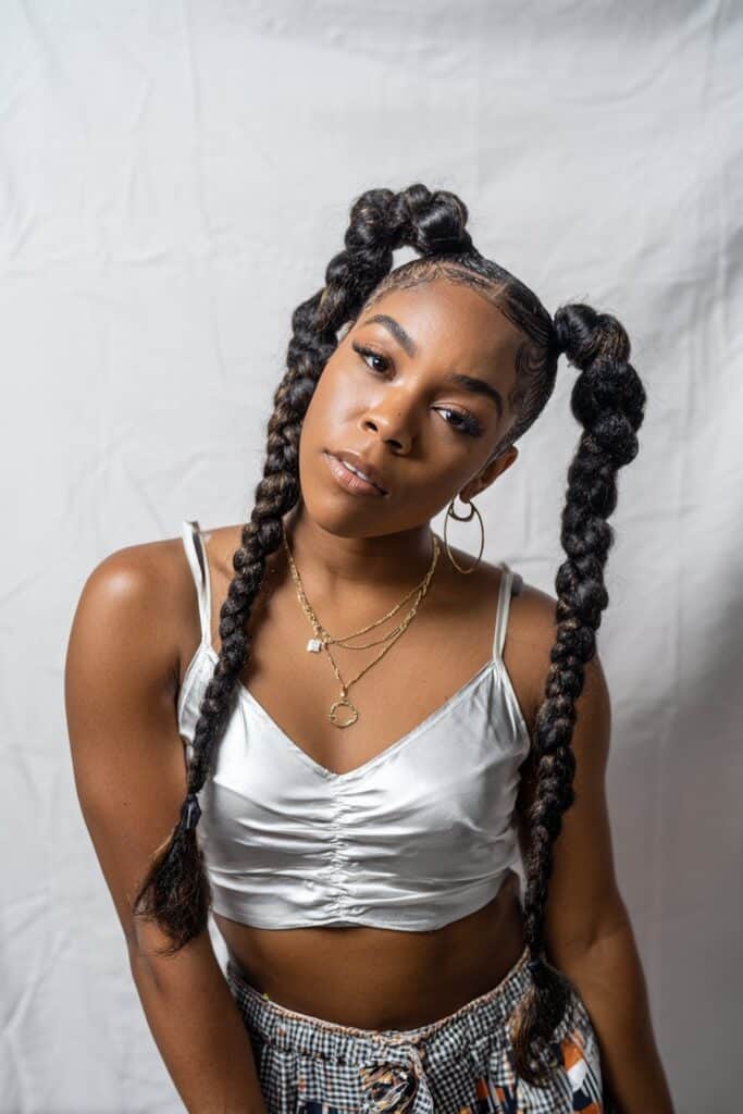 Playful portrait of American Black woman with two thick braids styled into high ponytails against white backdrop.