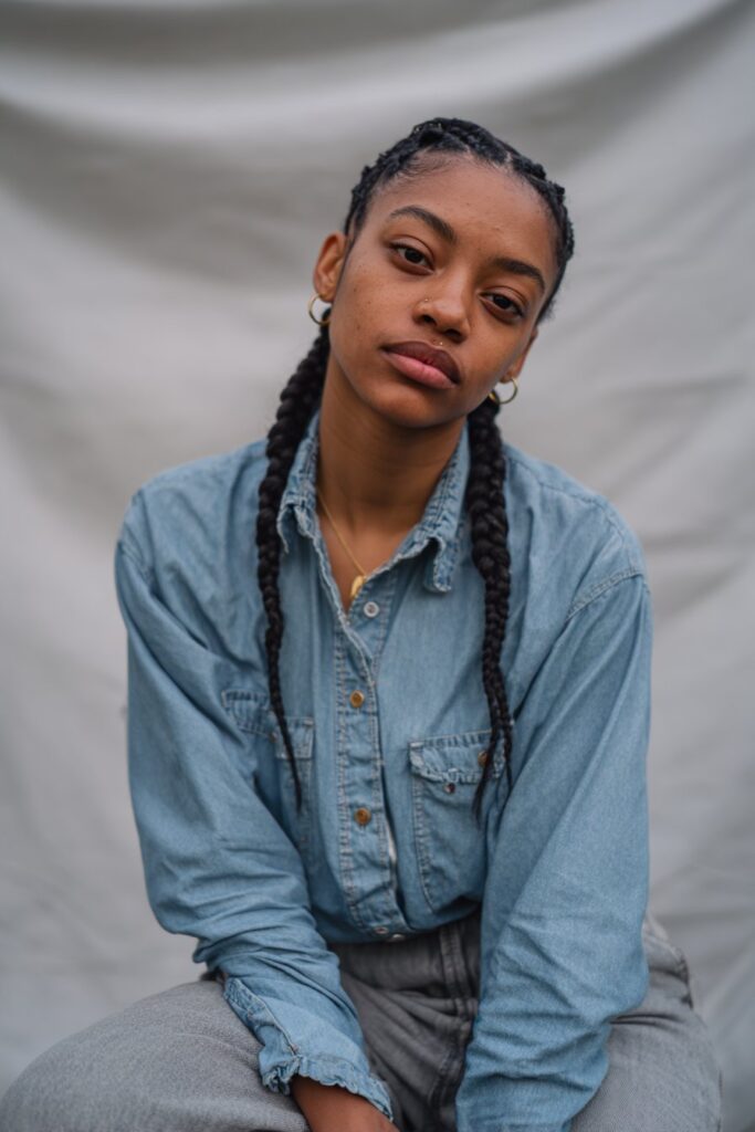 Playful portrait of American Black woman with double Dutch braid pigtails wearing casual denim against gray fabric backdrop.