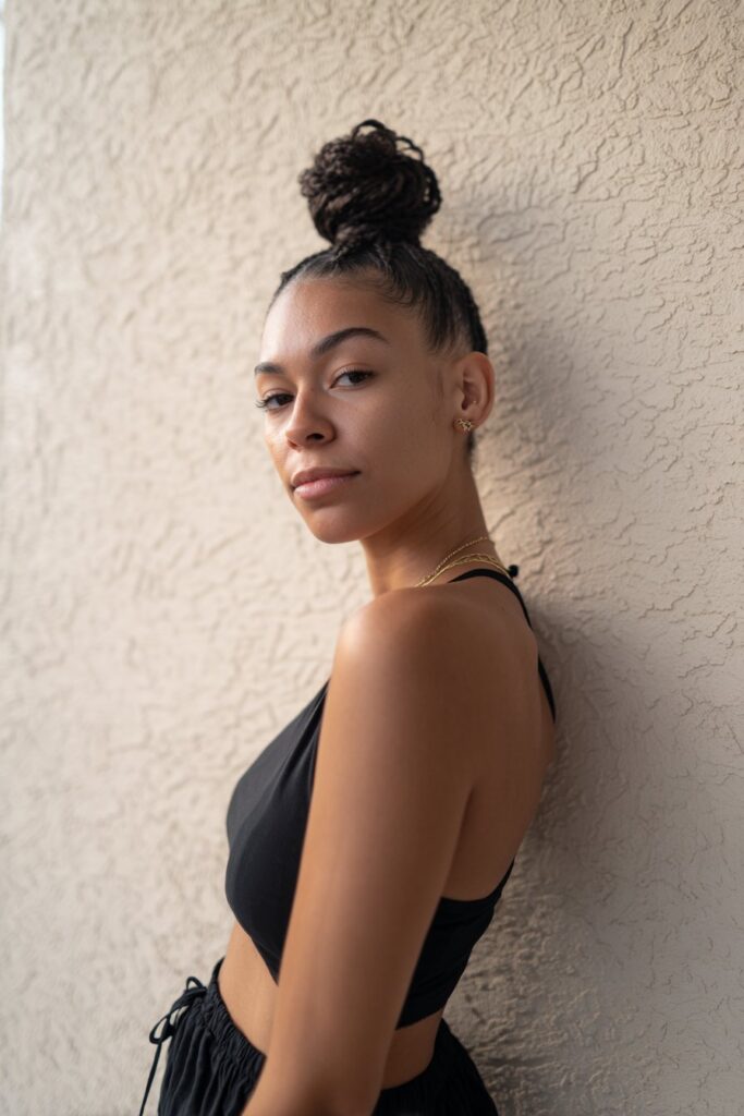 American woman with French braid top knot, confident pose, subtle glowy makeup against textured plaster background.
