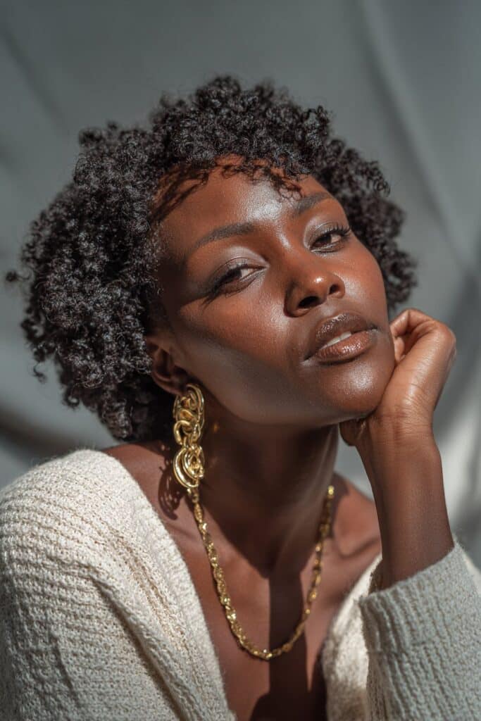 Beautiful Black woman with luxurious crochet curls, soft expression, gold accessories, gray fabric backdrop.