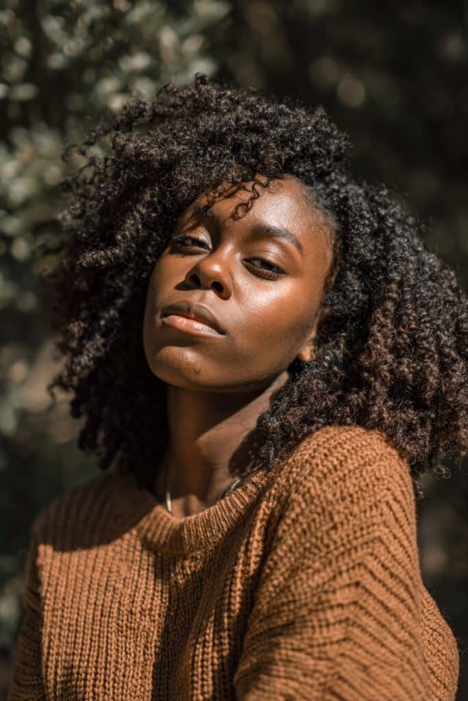 Confident Black woman with voluminous crochet curls, glowing skin, cozy knit top, natural forest backdrop.