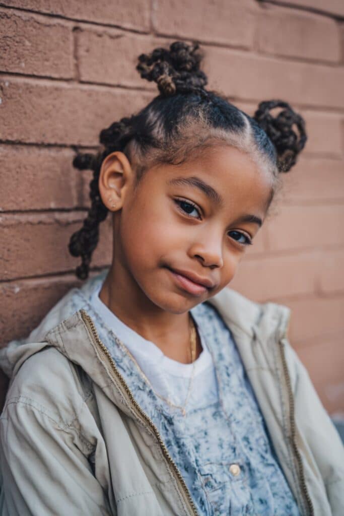 American girl with heart-shaped cornrows leading to double braided knots, wearing playful outfit, joyful expression against urban brick wall.