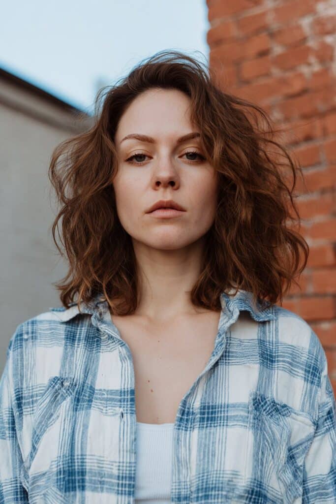 American woman with grunge bedhead hair inspired by Kurt Cobain, casual flannel shirt against urban brick wall backdrop.