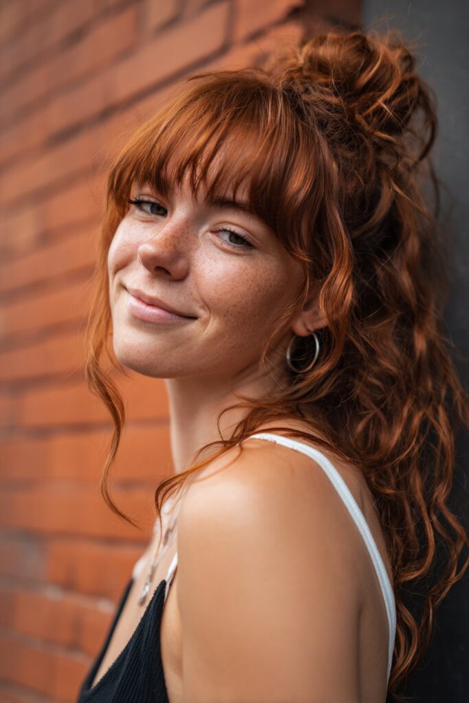 Playful portrait of American model with defined ringlet curls, full curtain bangs, casual outfit against brick wall.