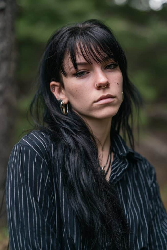 American female model with long black layered shag, curtain bangs, gothic eyeliner, striped emo shirt, forest scene background.