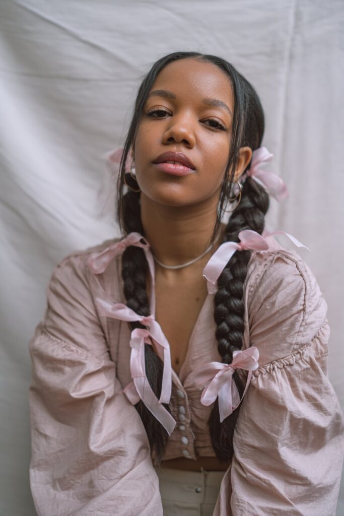Whimsical portrait of American woman with long fishtail pigtails tied with ribbons wearing pastel outfit against white backdrop.