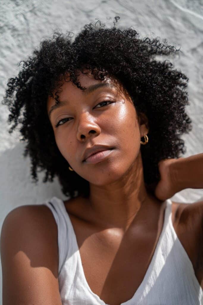 Black woman with full loose curl crochet afro, glowing skin, minimalist tank, textured plaster backdrop.